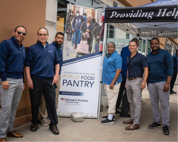 Six Mutual of America employees on a sidewalk posing in front of a sign with text “Catholic Charities of New York Pop-Up Food Pantry”
