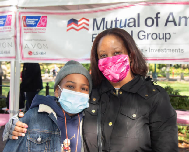 Masked child and mother standing outdoors in front of a Mutual of America sign at the American Cancer Society’s Making Strides Against Breast Cancer Walk